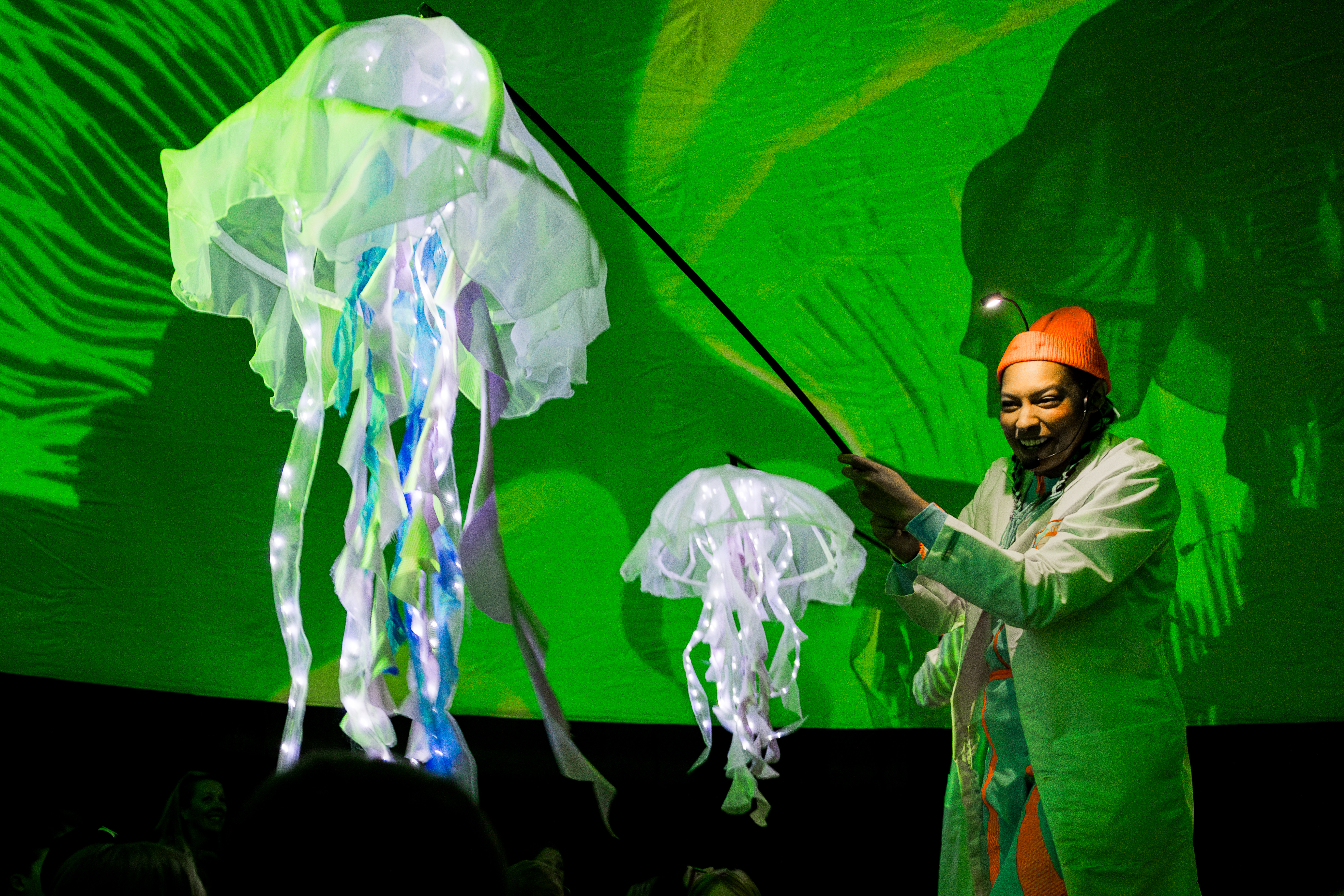 A woman operates a jellyfish puppet amidst a green projected background